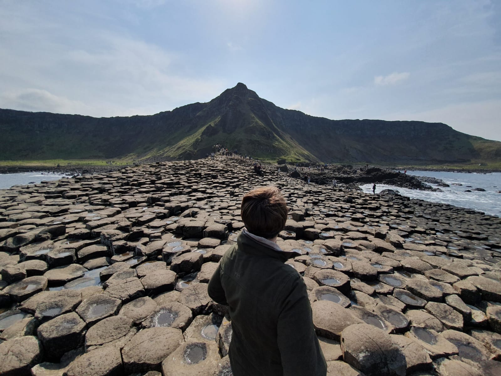 This is a photograph of Heather standing on the hexagonal rocks of the Giants Causeway. She is looking into the distance toward a mountain which sits at the centre of the picture.