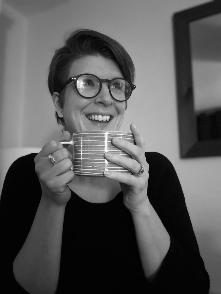 A black and white photograph of Heather looking off camera smiling and holding a cup of tea in both hands.